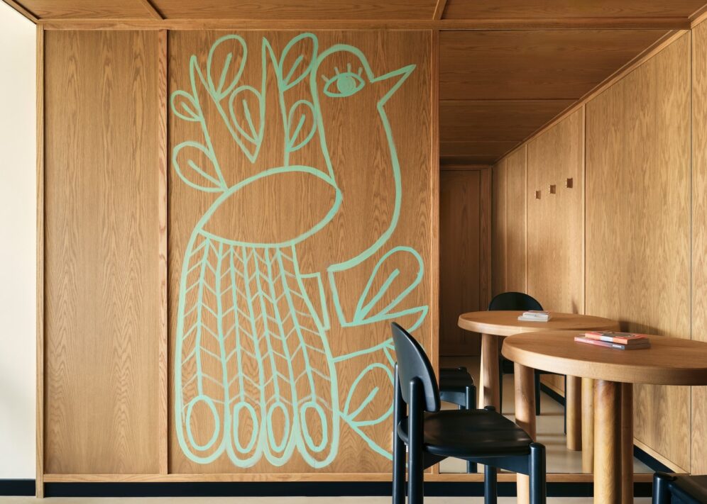A wooden room features an abstract teal bird design on the wall. The room has a warm and cozy ambiance, with a wooden table and black chairs.