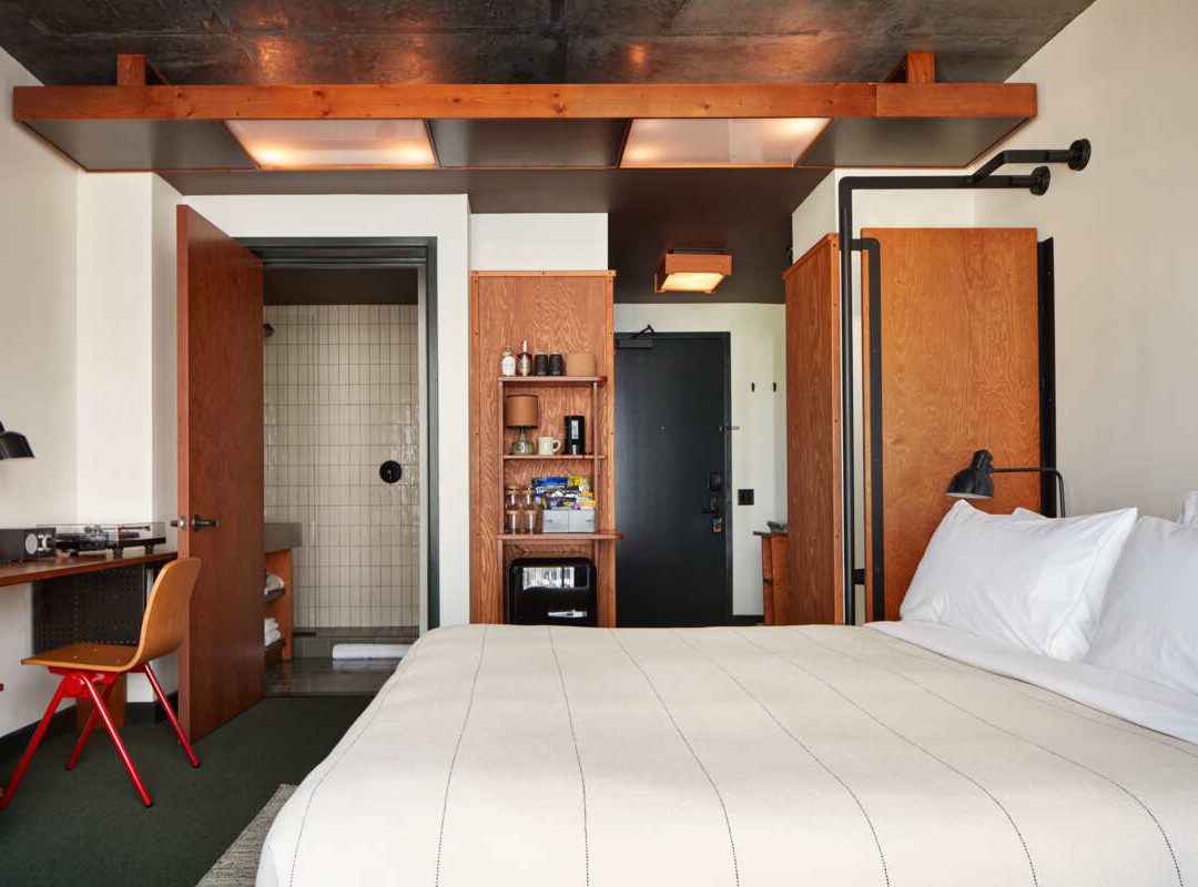 Modern hotel room with a neatly made bed, desk, open closet, and bathroom visible through a wooden door.