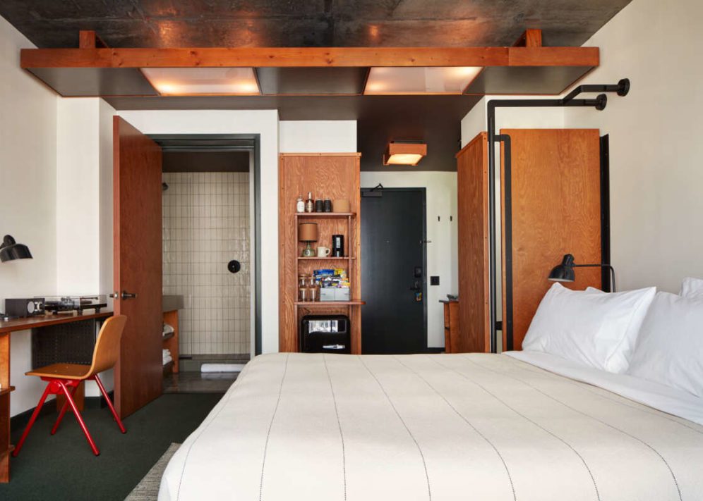 Modern hotel room with a neatly made bed, desk, open closet, and bathroom visible through a wooden door.