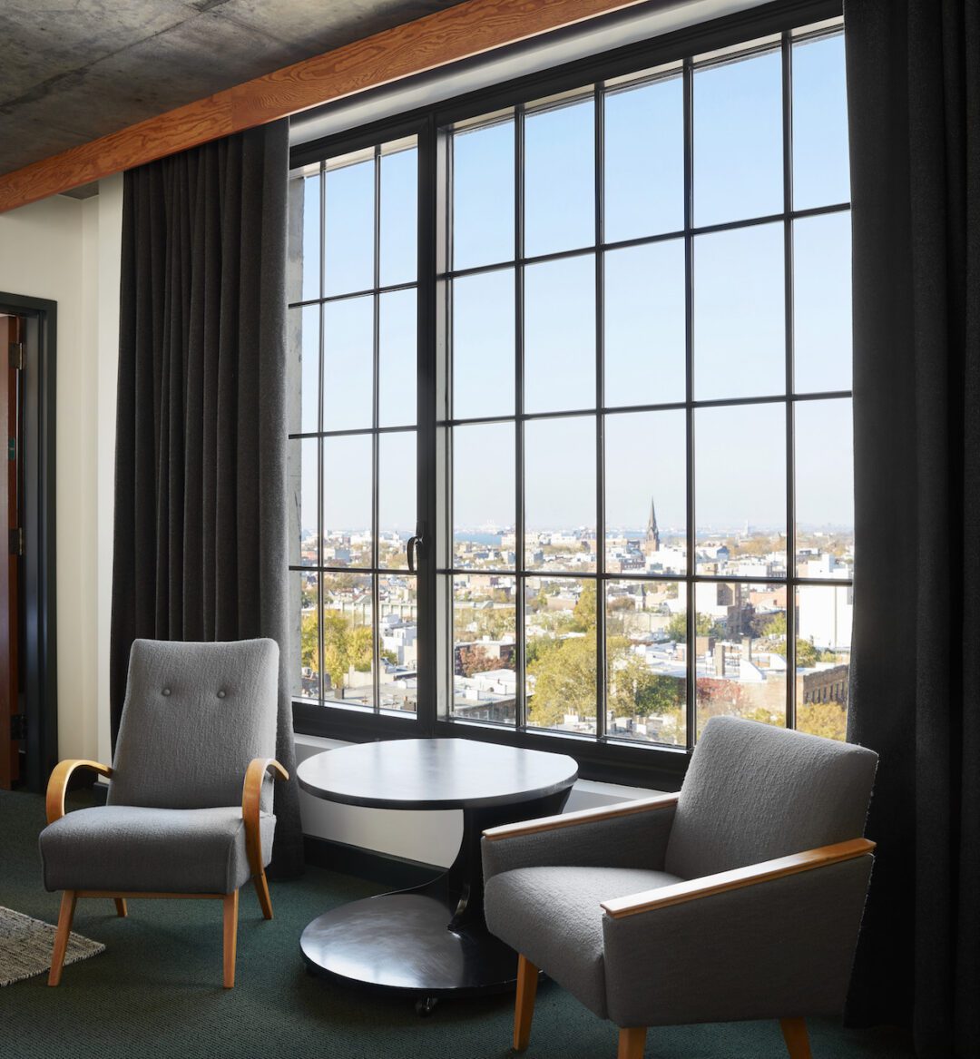 Two gray armchairs and a round table are positioned by a large window with black curtains, overlooking a cityscape.