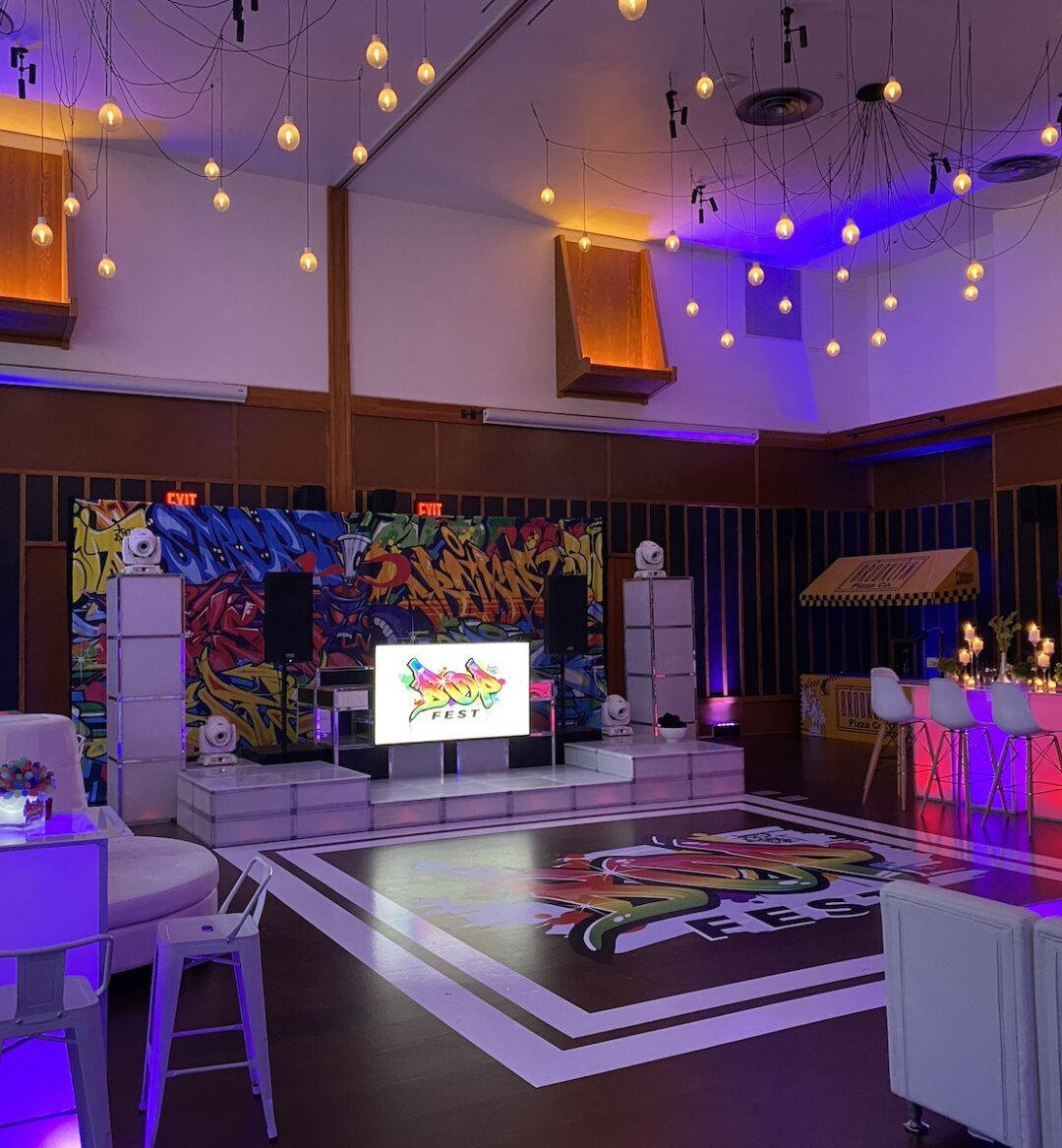 A decorated event space with colorful lights, graffiti-style art, lounge seating, and a central screen displaying Sushi Fest.