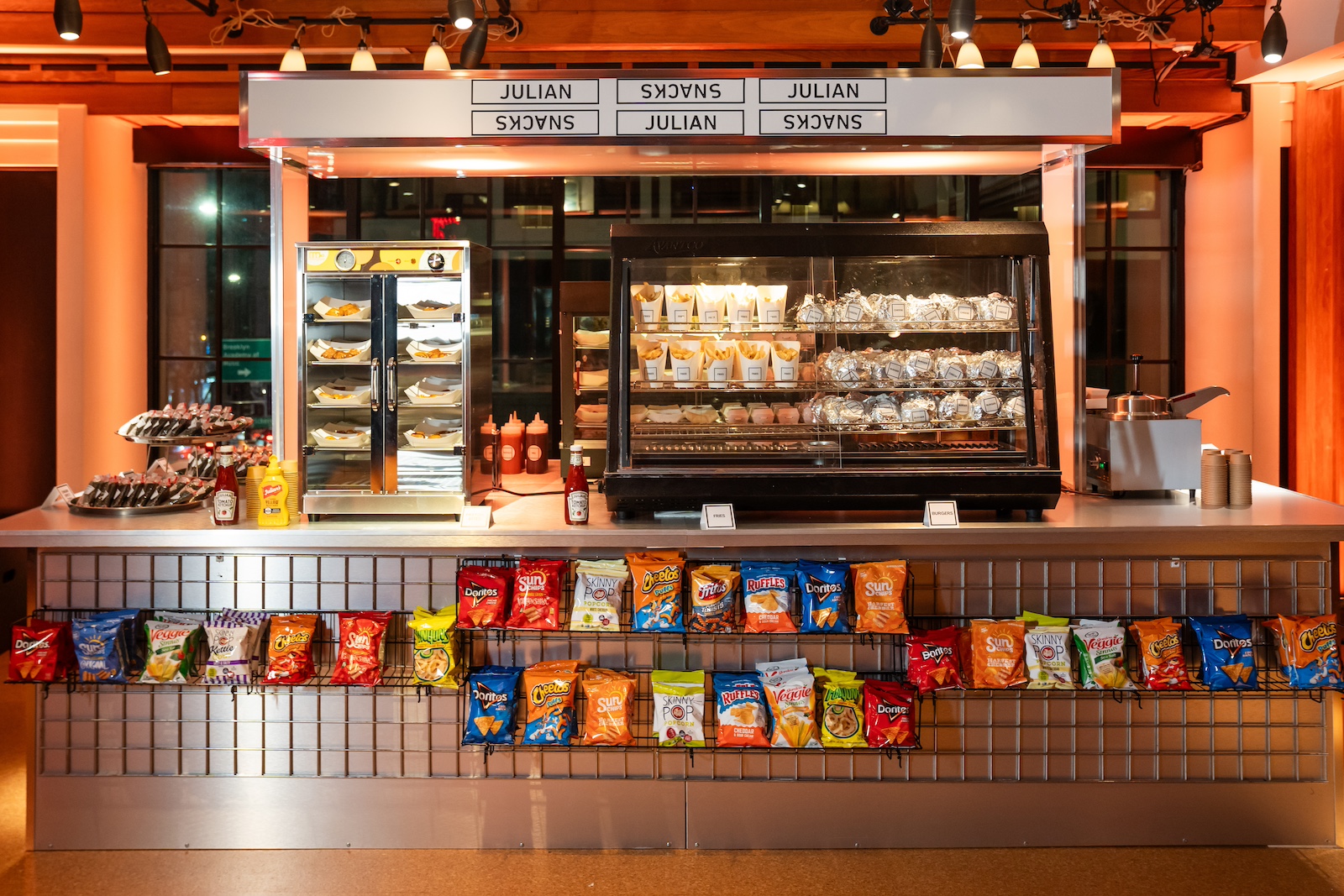 A snack stand displays packaged chips, pastries, and condiments under a sign reading Julian Snacks in a well-lit indoor space.