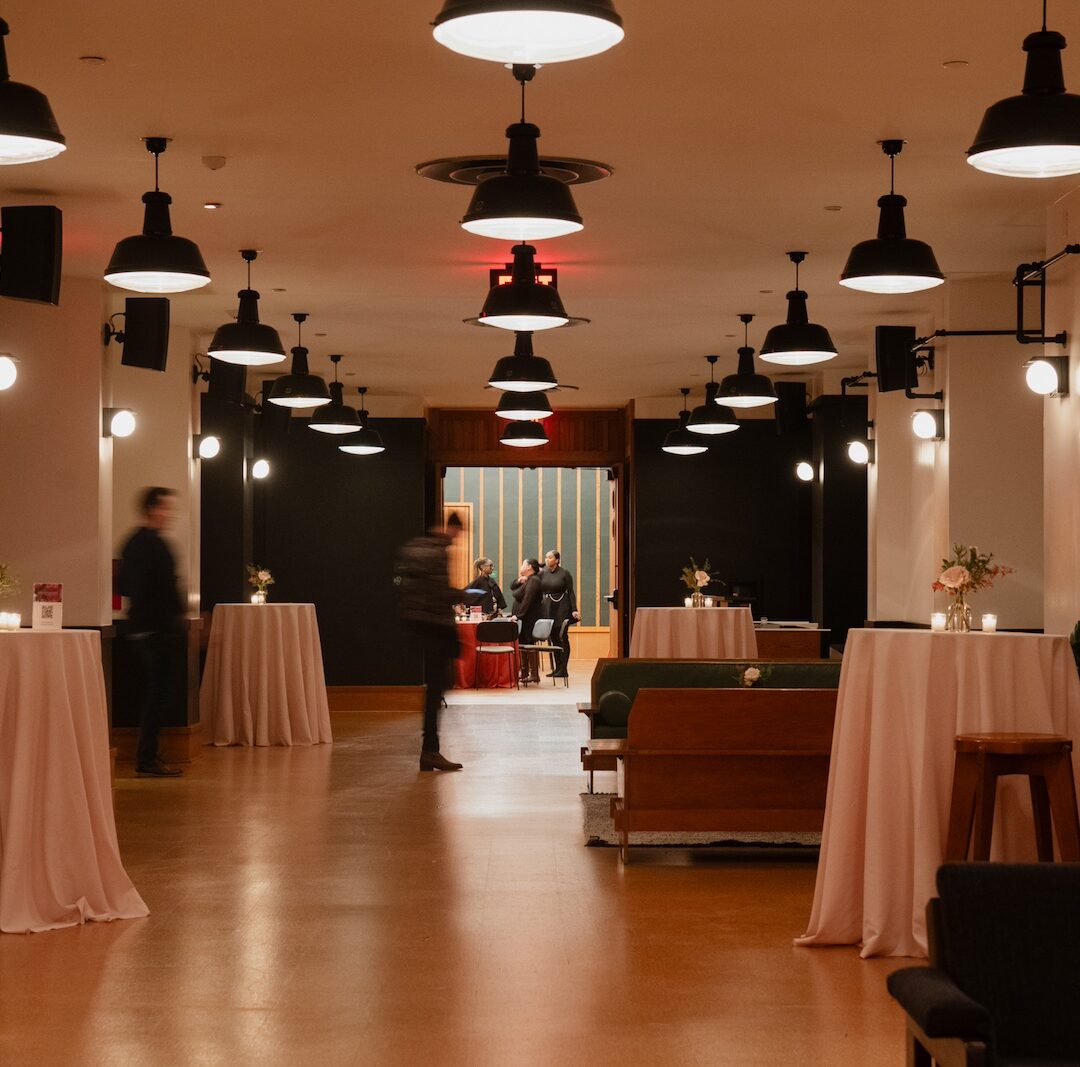 A dimly lit event space with high tables draped in pink cloth, benches, and a few people standing and sitting.