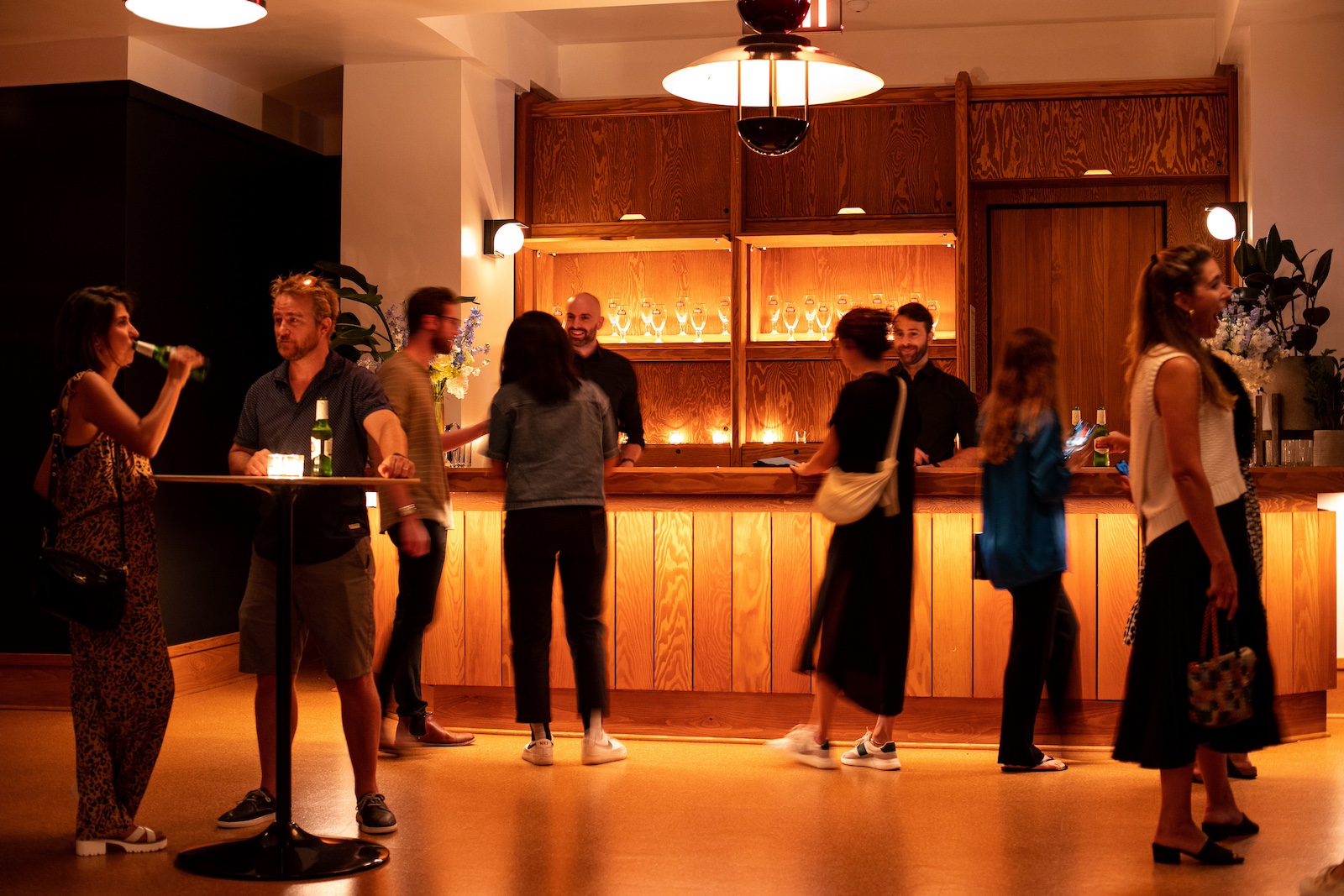 People stand and mingle near a wooden bar counter in a softly lit, modern indoor setting.