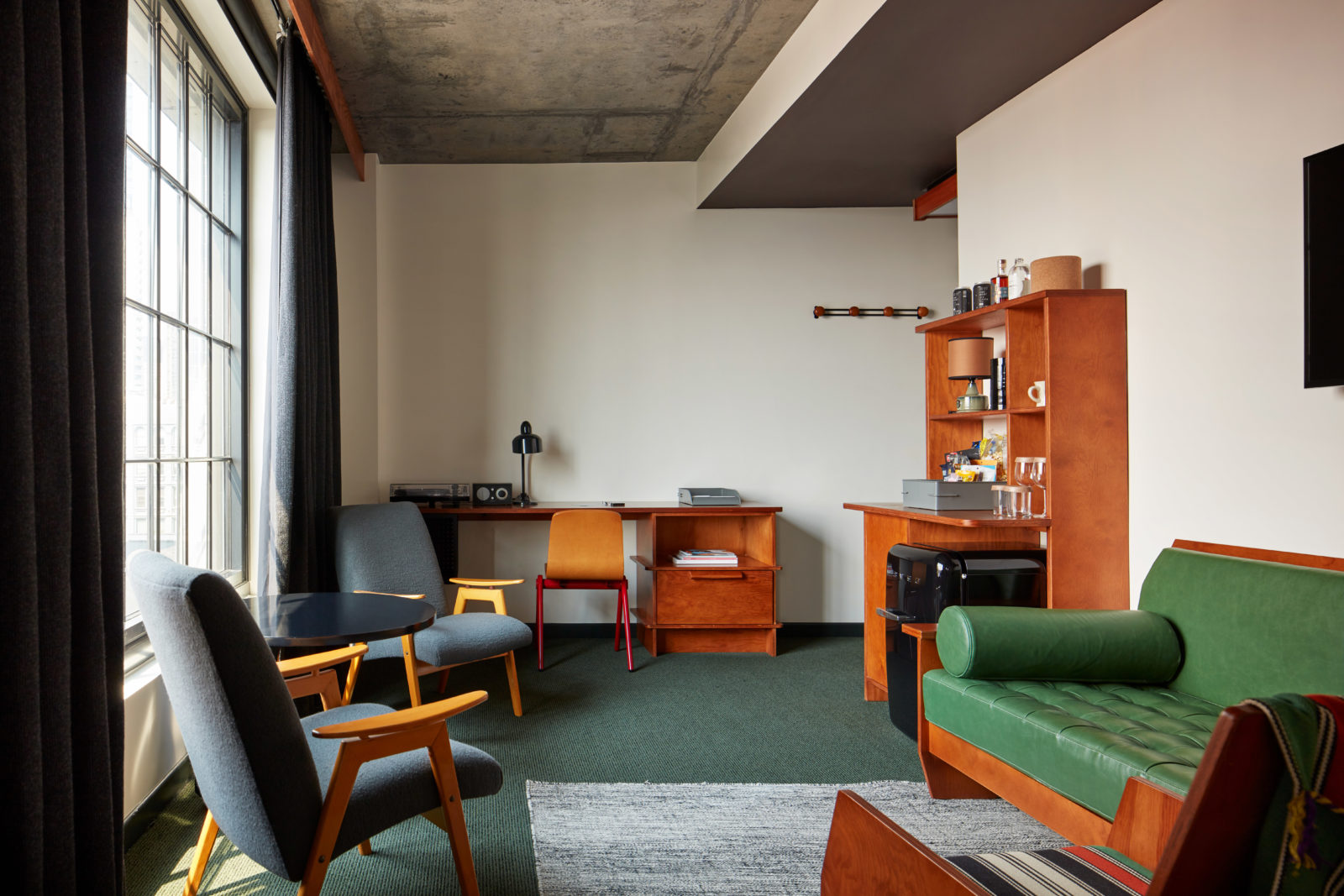 Hotel room with a green sofa, wooden furniture, desk, chairs, round table, and large window with dark curtains.