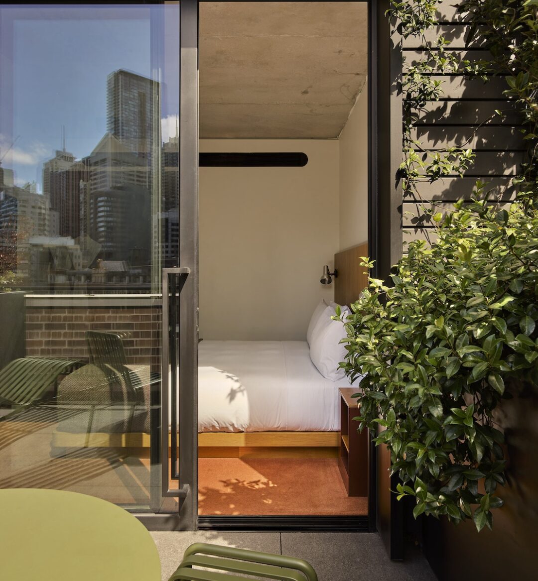 A hotel room with a bed seen through an open glass door to a small balcony with green chairs and city buildings in the background.