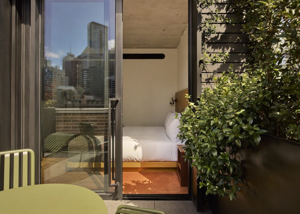 A hotel room with a bed seen through an open glass door to a small balcony with green chairs and city buildings in the background.