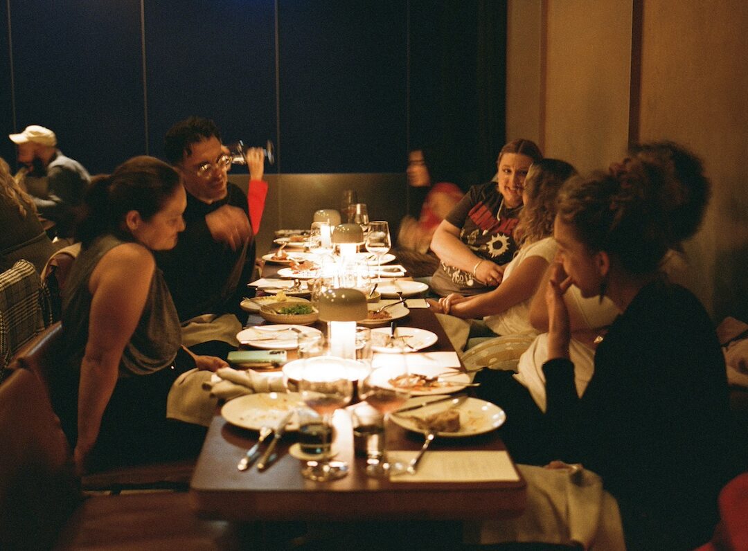 A group of people sit around a candlelit restaurant table, talking and eating dinner.
