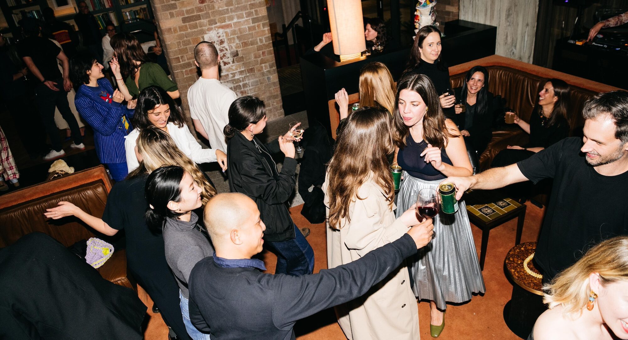 A group of people socialize, drink, and dance together in a dimly lit lounge with brown couches and brick walls.