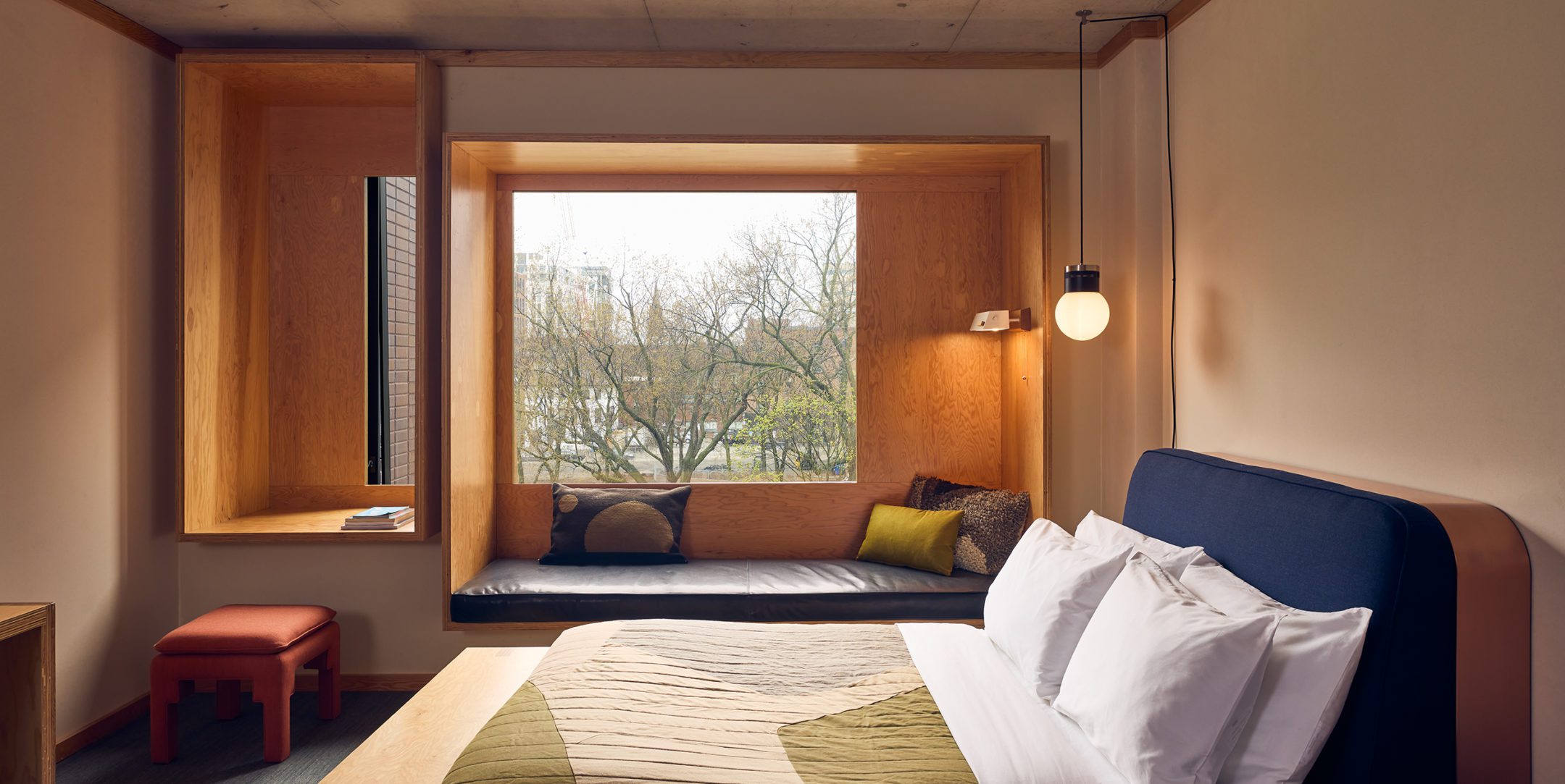 Modern hotel room with a bed, large window seat, pillows, and a view of trees outside. A pendant light hangs by the bed.