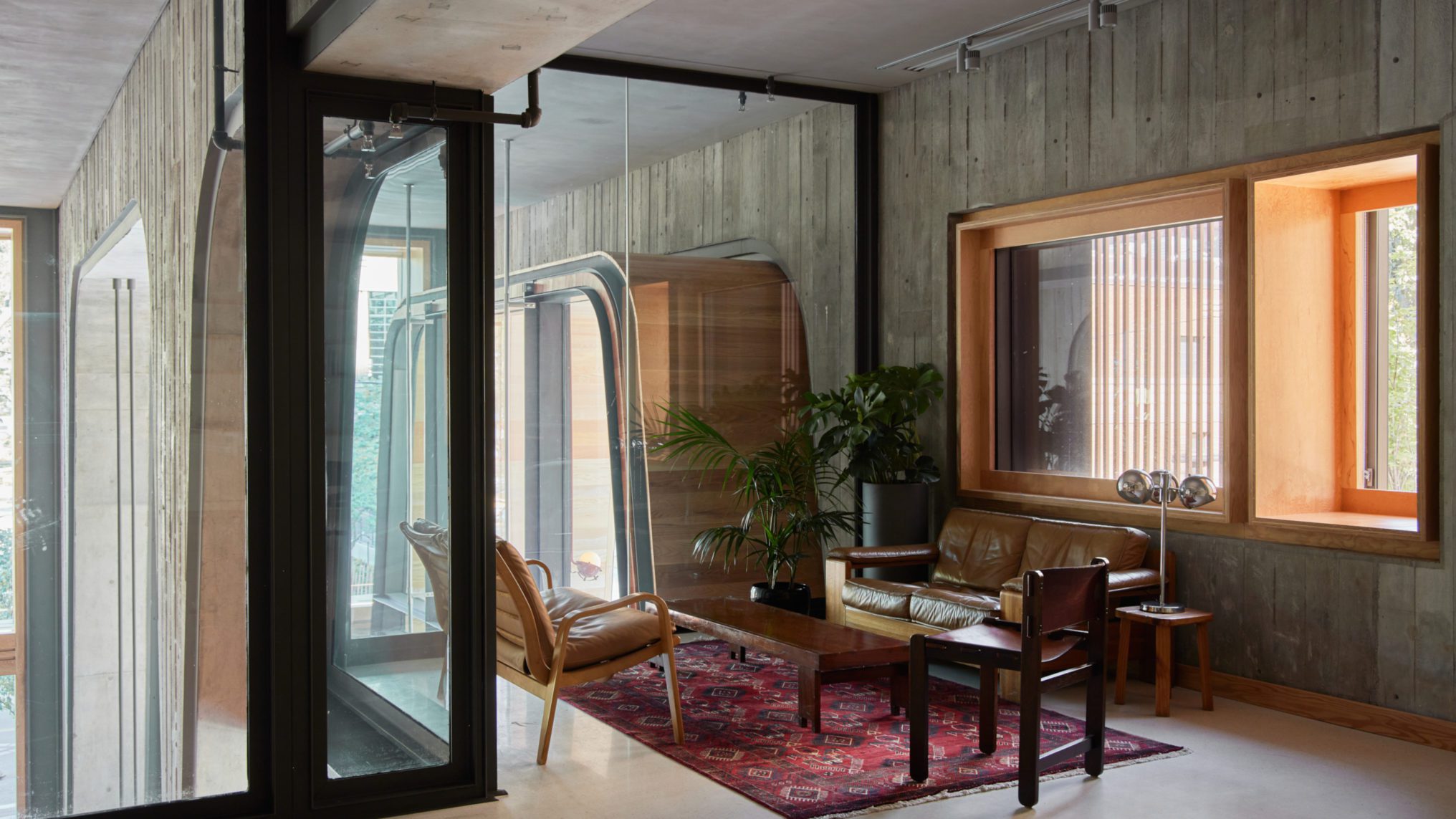Modern lounge with leather seating, wooden furniture, potted plants, large windows, and a red patterned rug on a concrete floor.