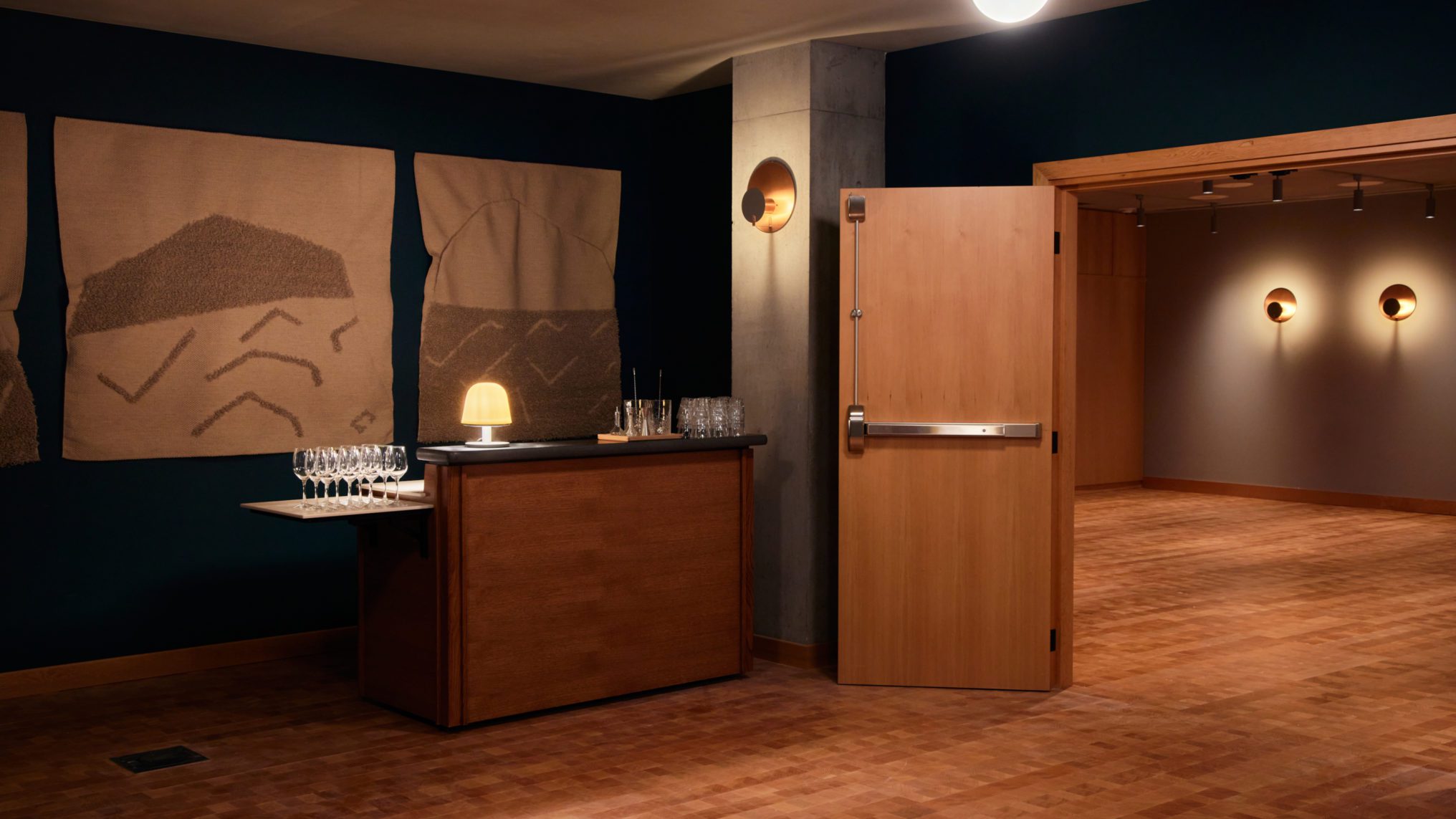 A dimly lit room with wall art, a small table set with glasses, and an open wooden door leading to another space.