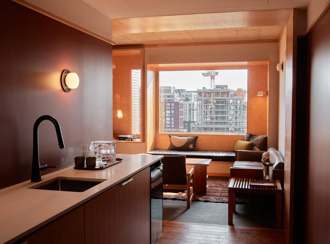 Modern hotel room with kitchenette, seating area by large window, city buildings visible outside, warm wood tones throughout.